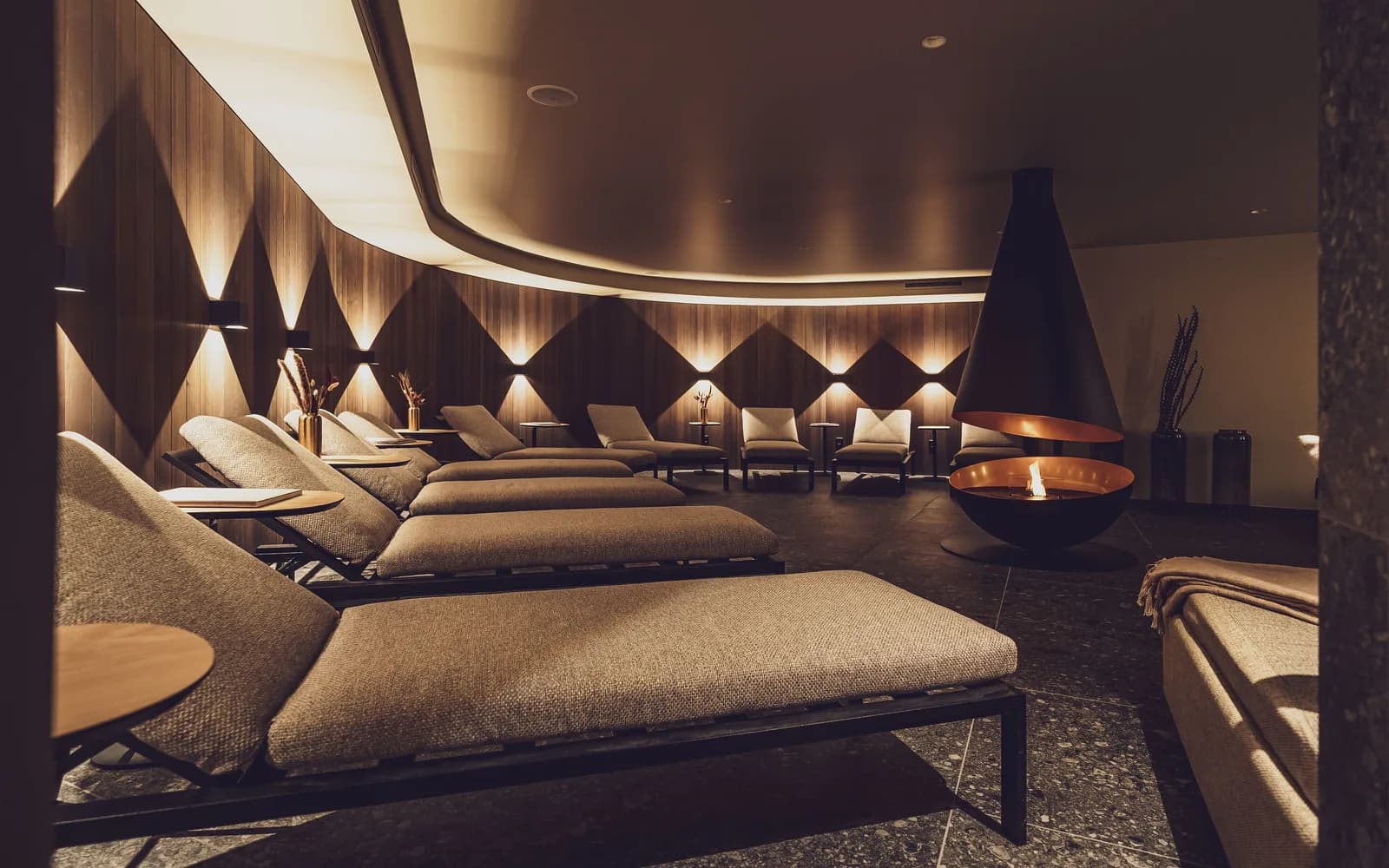 A softly lit spa relaxation lounge in a Davos hotel, with padded loungers arranged around a suspended fireplace.