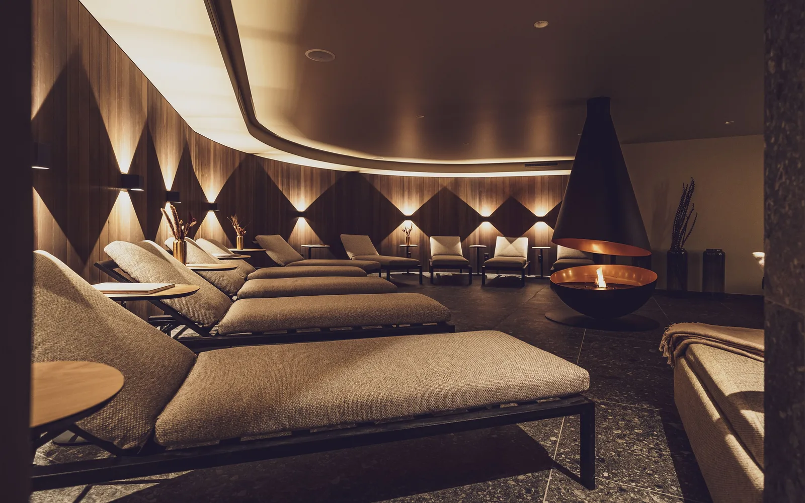 A softly lit spa relaxation lounge in a Davos hotel, with padded loungers arranged around a suspended fireplace.